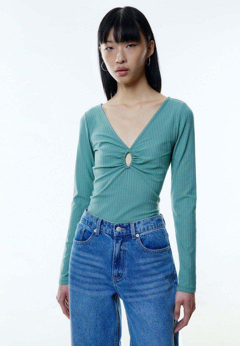 Mintgroene long-sleeve top met een V-hals en een keyhole-detail aan de voorkant, met ribbelstructuur. Gecombineerd met hooggetailleerde blauwe denim jeans.