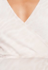 Κοντινό πλάνο ενός λευκού γυαλένιου ρούχου με V λαιμόκοψη, το οποίο φοριέται και αποκαλύπτει δέρμα μέσα από το λεπτό υλικό και την απαλή πτώση.