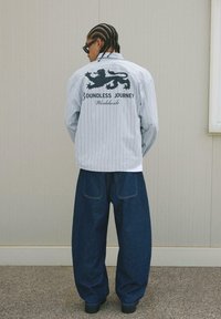 Camicia oversize a righe blu chiaro con grafica di un leone nero e testo sulla schiena, abbinata a pantaloni in denim scuri e larghi, indossata con scarpe nere.