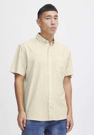 Short-sleeve, light beige button-up shirt with a chest pocket, made of a smooth fabric. Model wears it with blue jeans.