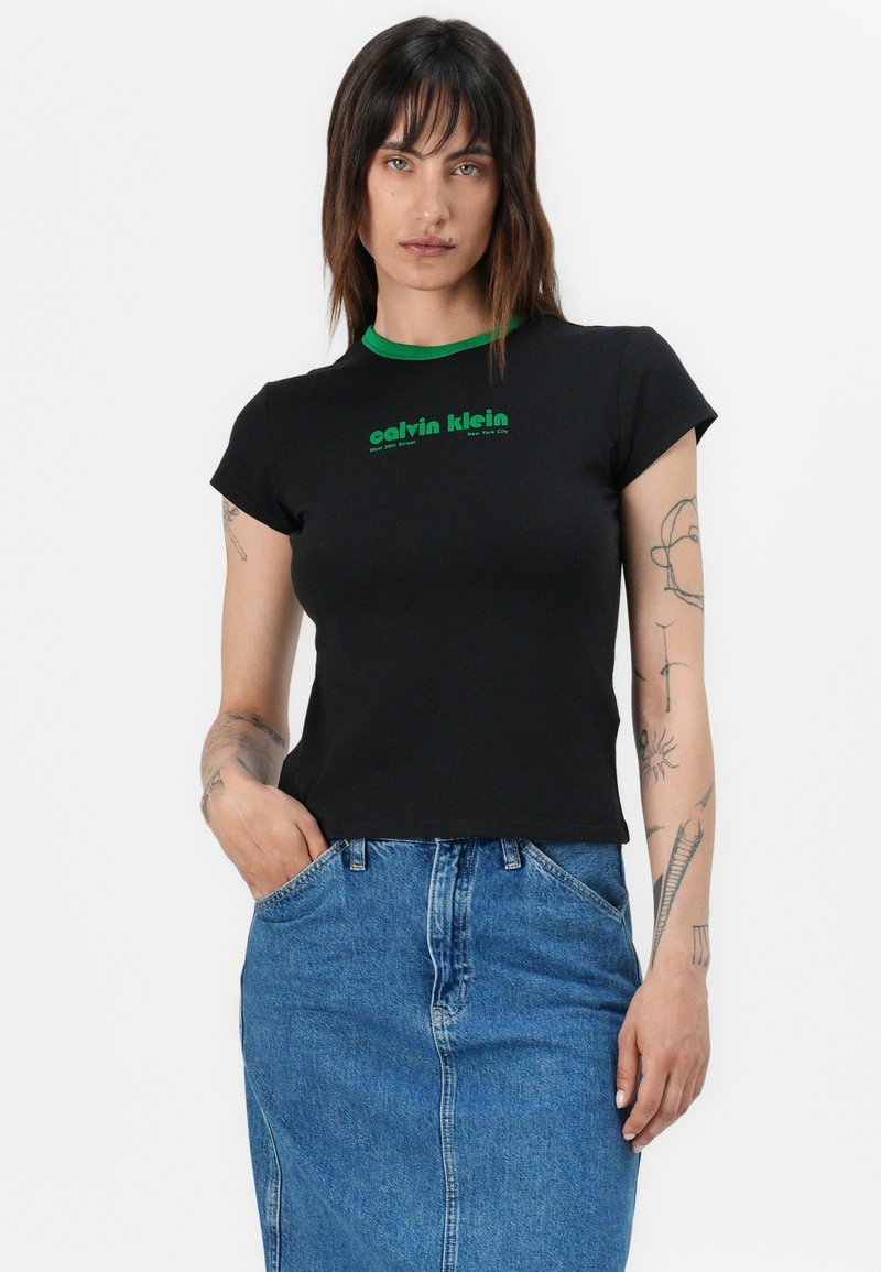 Schwarzes T-Shirt mit kurzen Ärmeln und grünem Kragen, mit "Calvin Klein" in grüner Schrift. Getragen mit einem blauen Jeansrock, der einen figurbetonten Stil zeigt.