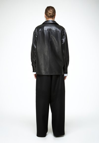 Hátulnézet, laza fekete bőrkabátot és széles szárú fekete nadrágot viselő, kontyba kötött hajú személy fehér háttér előtt.