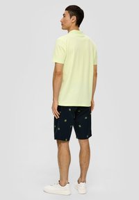 Helles Gelb Polohemd kombiniert mit marineblauen Shorts, die grüne Palmenmuster aufweisen. Das Modell trägt weiße Sneakers.