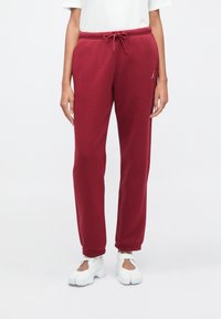 Άτομο που φοράει μπουρντό κόκκινες φόρμες με κορδόνι και μικρό λογότυπο στο αριστερό μηρό, καθώς και λευκά παπούτσια με κοψίματα, στέκεται σε λευκό φόντο.