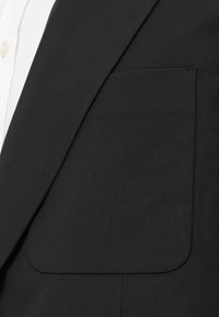 Tissu de costume noir avec une texture lisse ; présente une grande poche carrée sur le côté gauche, associée à une chemise blanche en dessous.