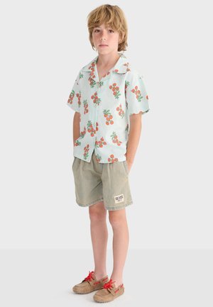 Garçon les mains dans les poches, portant une chemise à manches courtes claire avec un imprimé de fruits orange, un short beige et des mocassins marron à lacets rouges.