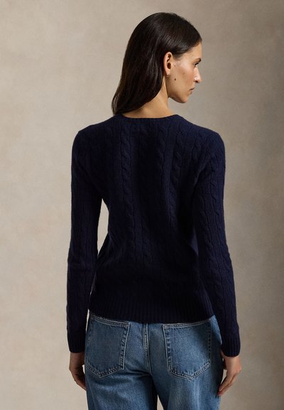 Femme aux cheveux foncés portant un pull torsadé bleu marine et un jean bleu, debout et tournée de dos sur un fond neutre.