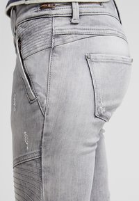 Gros plan sur un jean en denim gris clair avec des coutures texturées, un détail de fermeture éclair et des zones usées sur la poche arrière et la cuisse.
