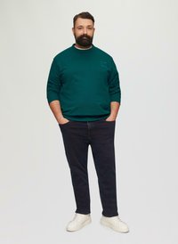 Sweatshirt vert à col rond et logo sur la poitrine, associé à un jean foncé et des baskets blanches. Texture lisse dans l'ensemble.