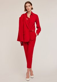 Rode maatpak met een overhemd met knopen en bijpassende blazer. Slim fit broek en witte puntige hakken; gladde stof.