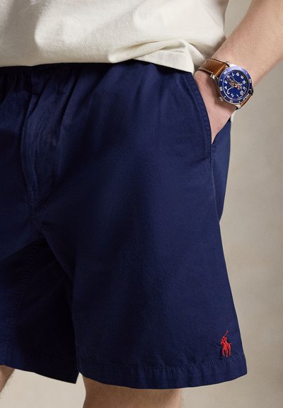 Osoba nosi tmavomodré šortky s červeným logom a na ľavom zápästí modré hodinky s hnedým koženým remienkom.