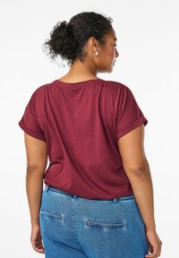 Ženska s kodrastimi lasmi z dvignjenimi lasmi, ki nosi srebrne uhane, maroon majico z zavihanimi rokavi in svetloblujši kavbojke, obrnjena bočno proti beli ozadju.