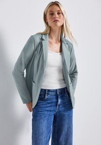 Světle modrý blazer se šitým designem, s klopou, dvěma bočními kapsami a hladkým povrchem, nošený přes bílé tričko a modré džíny.
