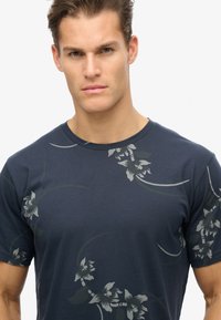 Camiseta de manga corta de color azul marino, confeccionada en una tela suave, que presenta un patrón floral en tonos grises y verdosos apagados en el pecho y los hombros.