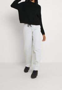Pull noir texturé avec un col montant, associé à un jean large gris clair et des baskets noires. La tenue présente une coupe décontractée et confortable.