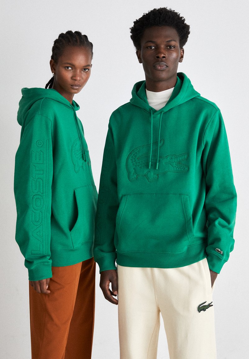 Sudaderas verdes fabricadas con material suave. Presenta un logo de cocodrilo en relieve en la parte delantera y el nombre de la marca a lo largo de la manga. Ajuste casual.