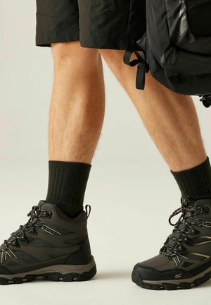 Homme portant des bottes de randonnée noires, des chaussettes noires et un short noir, portant un sac à dos noir, debout sur un fond clair.