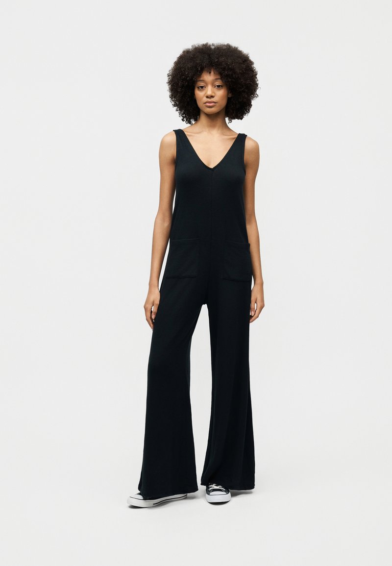 Femme aux cheveux bouclés portant une combinaison noire sans manches à jambes larges et des baskets noir et blanc, debout devant un fond gris uni.