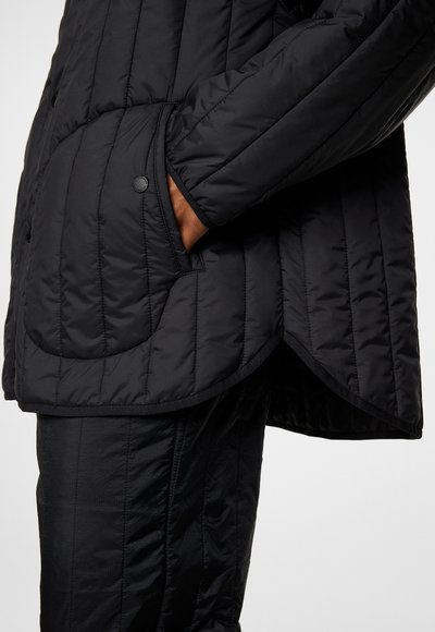 Svart vadderad jacka med diagonala sömmar, med rundad nederkant och sidfickor. Tillverkad av ett slätt, matt material med tryckknappar.
