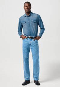 Camisa de mezclilla en azul mediano con dos bolsillos en el pecho, yugo puntiagudo y costuras en contraste. Combinada con jeans azul claro y botas negras de tacón.