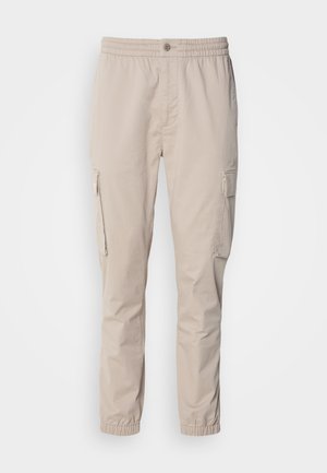 Beige cargo pants with an elastic waistband, button closure, and side pockets. Smooth fabric with tapered legs and elastic cuffs.