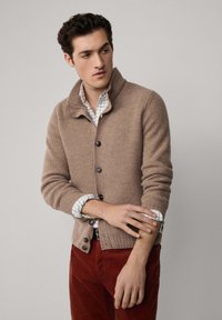 Béžový pletený kardigán s vysokým golierom, s tmavými gombíkmi a rebrovanými manžetami, nosený cez modro-bielu kockovanú košeľu, doplnený červenými nohavicami.