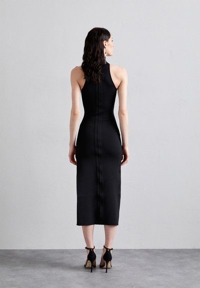 Abito midi nero aderente con collo alto, dettaglio della zip sul retro e tessuto liscio. Modello abbinato a scarpe nere con tacco alto, che mette in risalto una silhouette elegante.