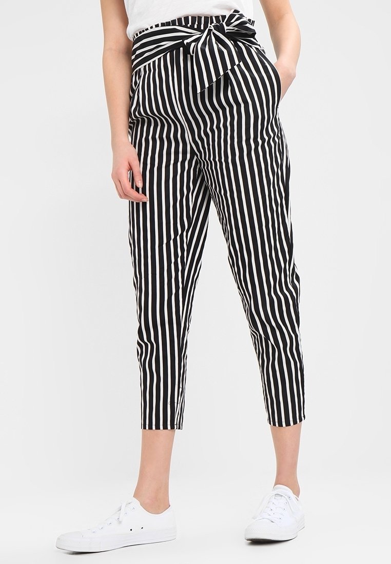 Schwarz-weiße gestreifte Hosen mit hoher Taille, Schleifendetail, schmal zulaufenden Beinen und cropped Länge, kombiniert mit weißen Sneakern.