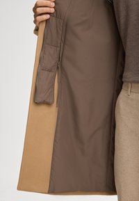 Cappotto beige con una morbida fodera interna marrone. Presenta una tasca con zip sulla parte interna sinistra e una superficie esterna morbida.