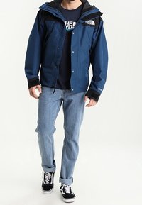 Modrá nepromokavá bunda s černými akcenty, nastavitelnou kapucí, kapsami na zip, v kombinaci s světle modrými džíny a černými teniskami.