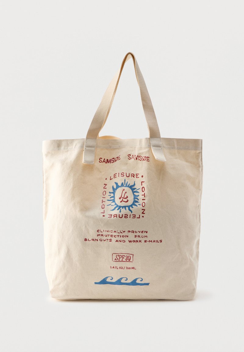 Sac fourre-tout en toile beige avec poignées beiges, graphiques rouges et bleus représentant un soleil et des vagues, et texte concernant une lotion de loisirs et une protection SPF 30.