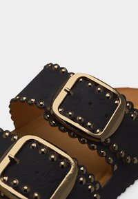 Μαύρα λουριά δερμάτινων σανδαλιών με χρυσές τετράγωνες αγκράφες και ταιριαστά χρυσά καρφιά κατά μήκος των κυματιστών ακμών.