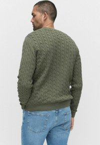 Grön kabelstickad tröja med ett texturerat mönster, rundad nacke och ribbad mudd, tillsammans med ljusblå jeans.