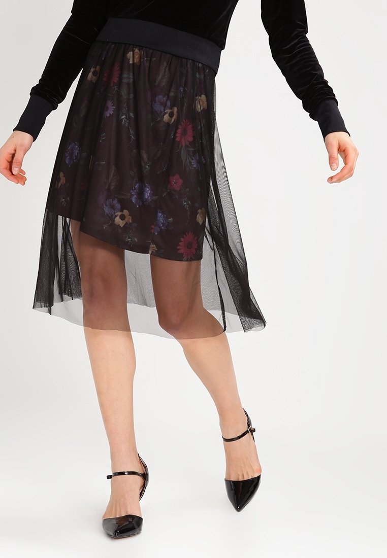 Femme portant une jupe noire florale transparente avec un jupon noir uni et des talons noirs à bride cheville à bout pointu, debout devant un fond blanc.