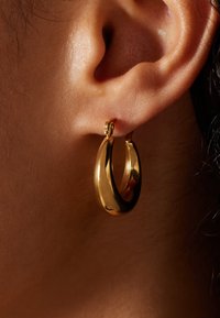 Gold hoop earring with a smooth, polished surface. The design is circular with a slightly elongated shape, hanging from a small clasp.