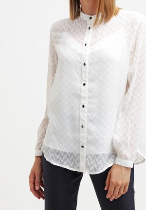 Weiße, strukturierte Bluse mit langen Ärmeln und schwarzen Knöpfen, getragen zu schwarzen Hosen, Modell von den Schultern bis zu den Hüften gezeigt.