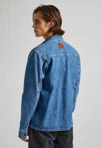 Denim jakna v srednje modri barvi s klasično obliko, rjav usnjen žep na zadnji strani in gumbi na manšetah. Ima rahlo teksturirano tkanino.