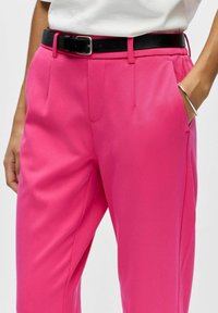 Pantalon rose vif avec une texture lisse, des jambes fuselées et des poches latérales, accompagné d'une ceinture en cuir noire pour le contraste.
