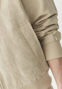 Beige bomberjakke i blødt, struktureret stof, med ribstrikkede manchetter og en enkelt brystlomme på venstre side. Casual, løs pasform.