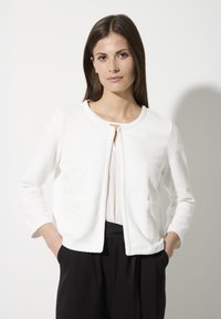 Weiße, strukturierte Jacke mit Rundhalsausschnitt, langen Ärmeln und zwei Fronttaschen, kombiniert mit einem hellen Oberteil und schwarzen Hosen.