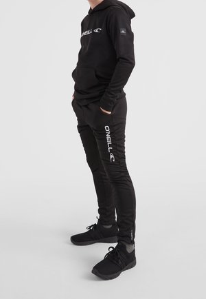 Zwarte hoodie met voorslaapzak en wit logo, gecombineerd met zwarte slim-fit broek met witte branding op het linkerbeen en zwarte sneakers.