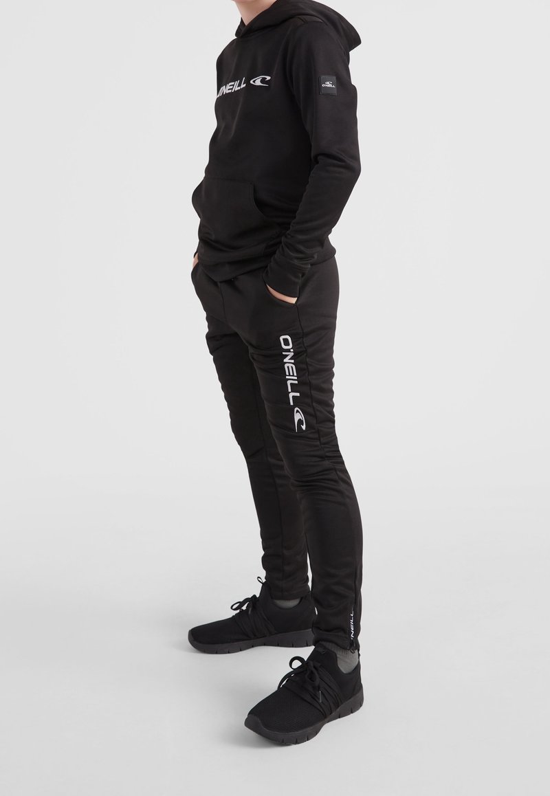 Sweat à capuche noir avec poche avant et logo blanc, assorti à un pantalon noir slim avec marquage blanc sur la jambe gauche et des baskets noires.