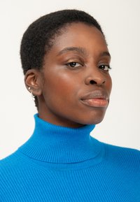 Maglione a collo alto blu a coste con un collo alto. Incarnato medio scuro, capelli corti e texturizzati, orecchini sottili e trucco naturale.