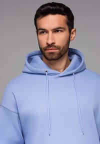 Hoodie bleu clair en tissu doux, avec un cordon de serrage en métal argenté, une capuche spacieuse et une coupe décontractée.