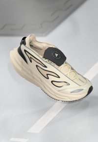 Zapatilla beige con suela chunky, diseño de malla, acentos negros y un parche con el logotipo. Presenta curvas dinámicas y superficies texturizadas.