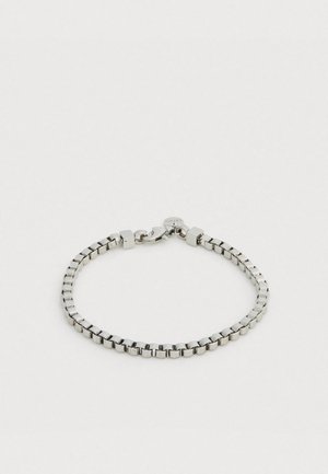 Srebrna metalowa bransoletka z kwadratowymi ogniwami o gładkim wykończeniu, zapinana na zapięcie typu karabińczyk i zawierająca małą, okrągłą zawieszkę z logo marki.