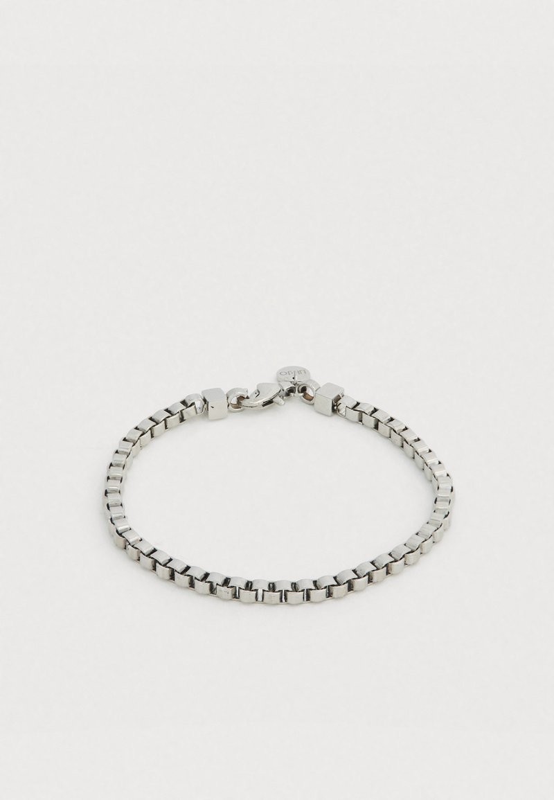 Brățară din metal argintiu cu zale pătrate cu finisaj neted, prinsă cu o închizătoare tip homar și o etichetă mică circulară cu marca.