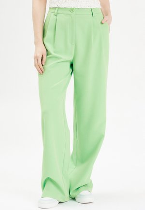 Groene wijde broek gemaakt van soepel stof, met een voorplichet, zijzakken en een knoopsluiting. Gekozen in combinatie met witte sneakers.