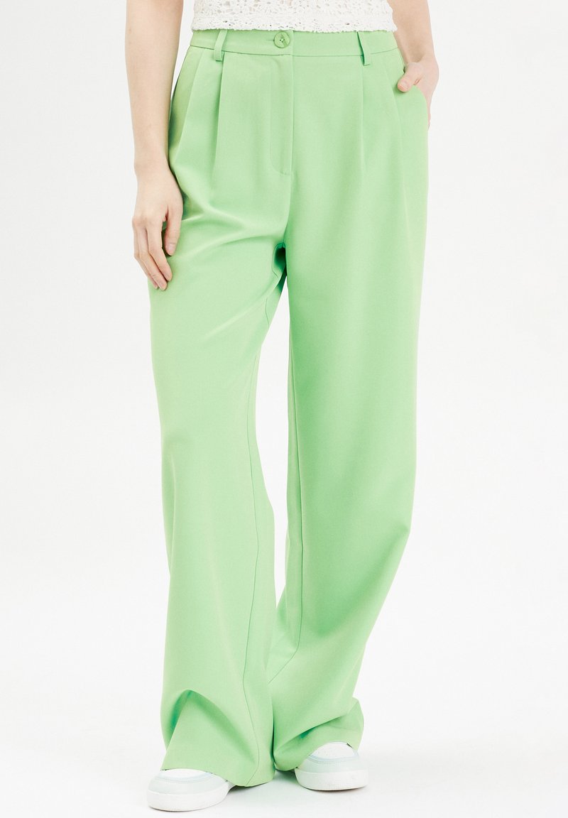 Groene wijde broek gemaakt van soepel stof, met een voorplichet, zijzakken en een knoopsluiting. Gekozen in combinatie met witte sneakers.