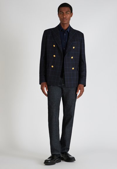 Man in een donkerblauwe geruite dubbelknoop blazer met gouden knopen, een overhemden in het donker, donkerdenim jeans en zwarte leren instappers, staand.